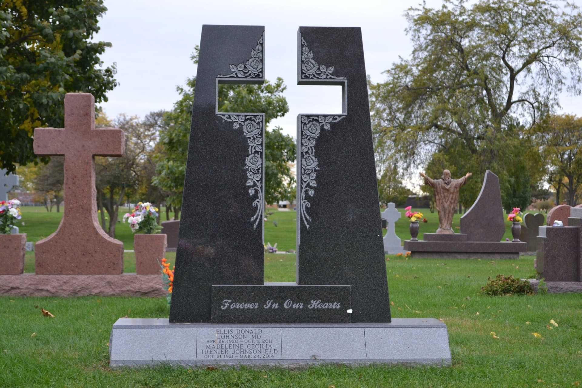 A black granite gravestone with a cross-shaped cutout, etched with floral designs, set in a green cemetery.