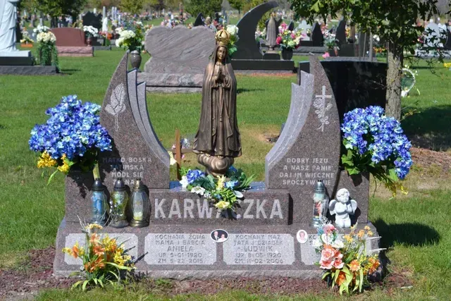 A brown granite headstone with a statue of the Virgin Mary, surrounded by floral arrangements, in a cemetery.