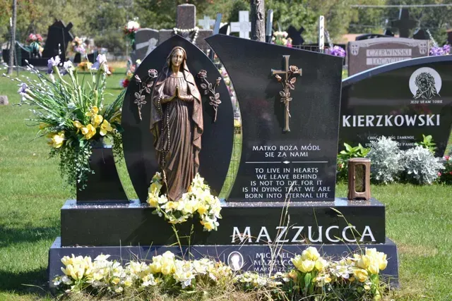 A black granite headstone with a bronze Virgin Mary statue, a cross, and yellow flowers in a cemetery, inscribed MAZZUCCA.