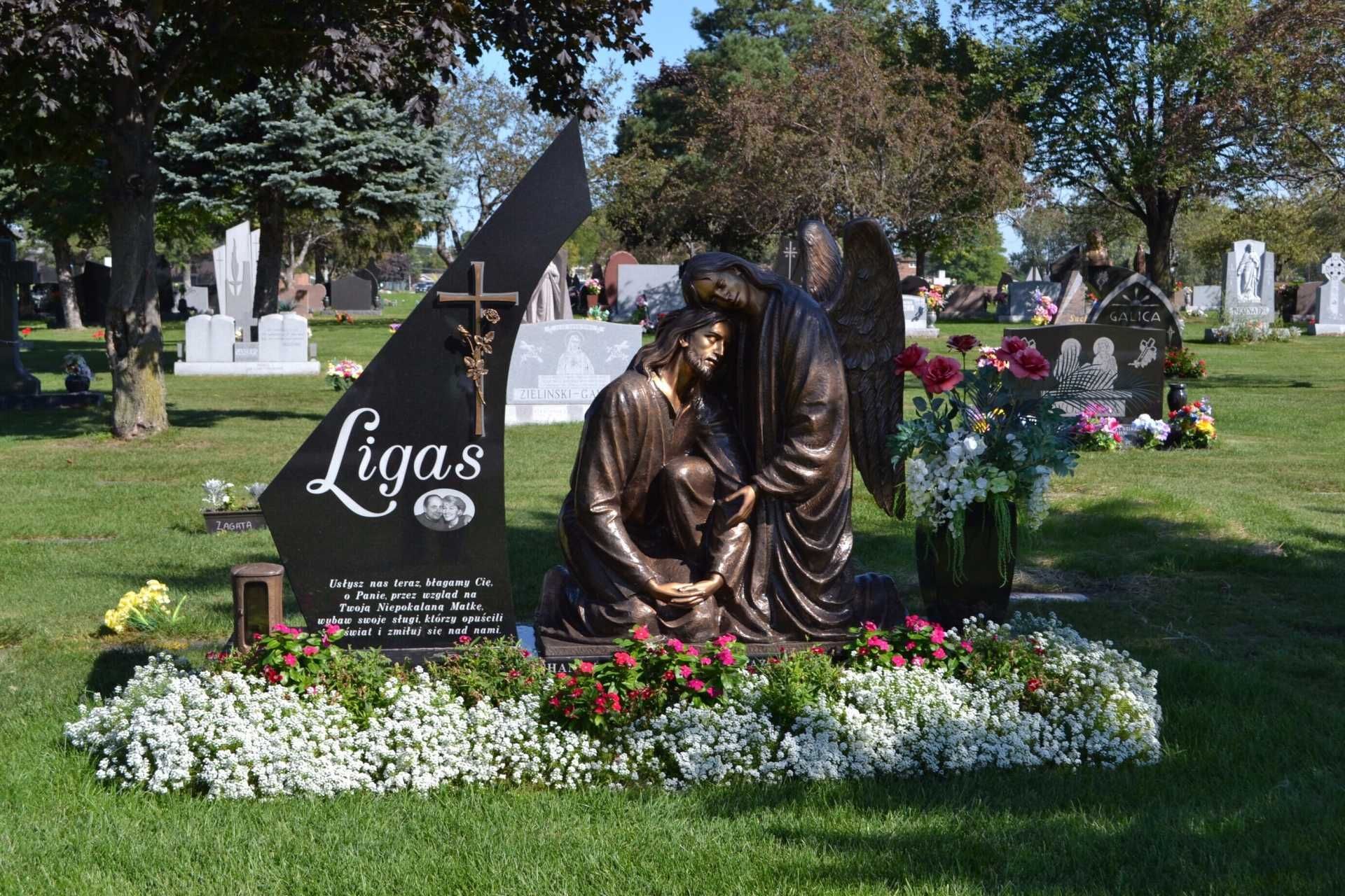Cemetery headstone with bronze angel statue kneeling beside a person. Colorful flowers surround the monument.