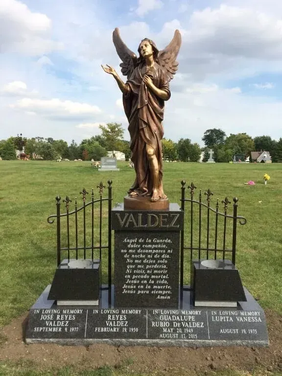 A bronze angel statue stands atop a black granite monument with inscriptions, framed by an iron fence in a cemetery.