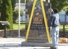 Person adjusting a large, dark stone memorial with yellow straps. Outdoor setting.