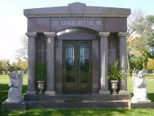 A dark stone mausoleum with a central door, decorative columns, and two angel statues in a grassy cemetery setting.
