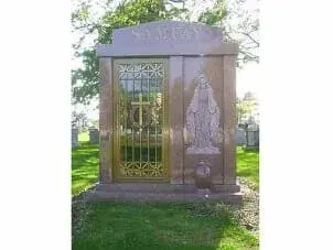 A granite outdoor mausoleum with a gold-framed glass door featuring a cross and a carved relief of a figure on the right.
