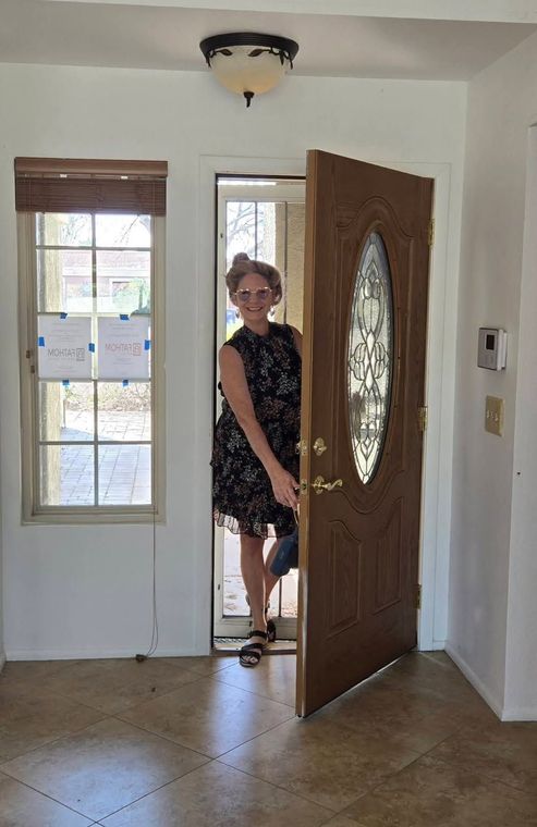 Woman standing in a doorway, partially behind an open wooden front door in a bright entryway.