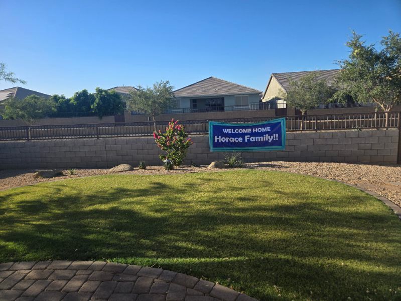 Front yard with a blue “Welcome Home” sign, grass lawn, stone wall, and suburban houses under a clear sky