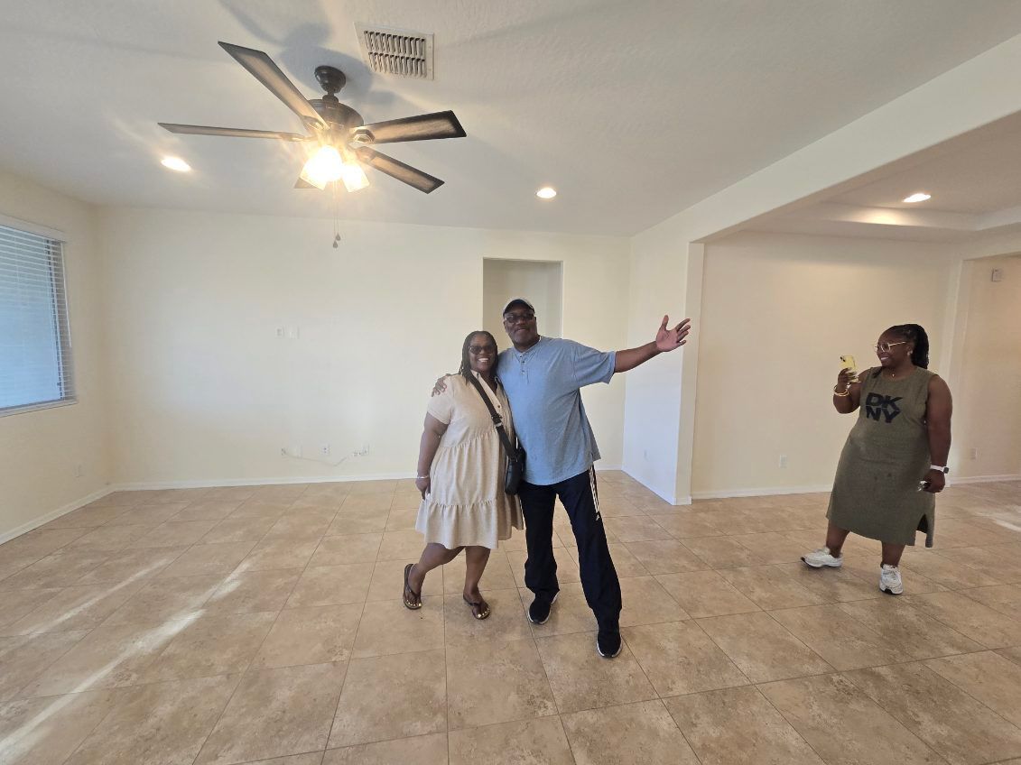Three people in an empty beige room, one posing with arms outstretched under a ceiling fan.