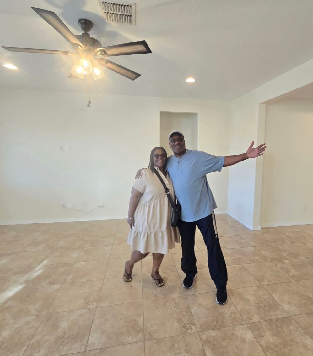 Two people posing in an empty room with tile floor, white walls, and a ceiling fan.