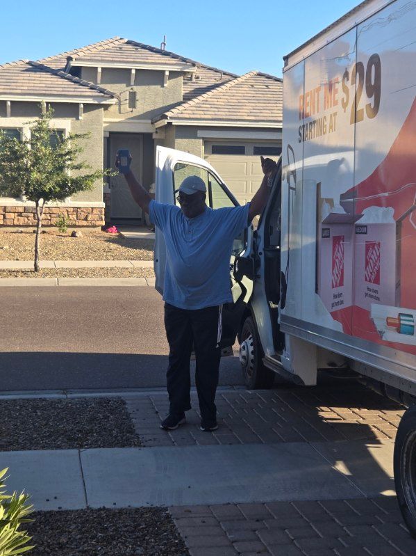 Person in blue hoodie stands beside a moving truck with arms raised in a suburban driveway.