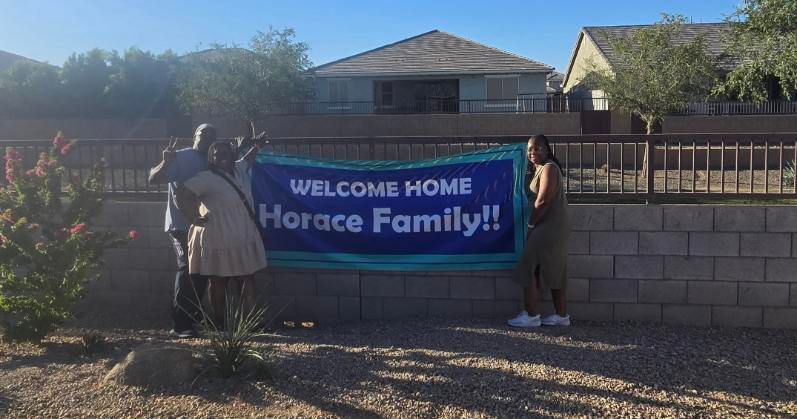 Two people hold a blue “WELCOME HOME Horace Family!” banner in a front yard.