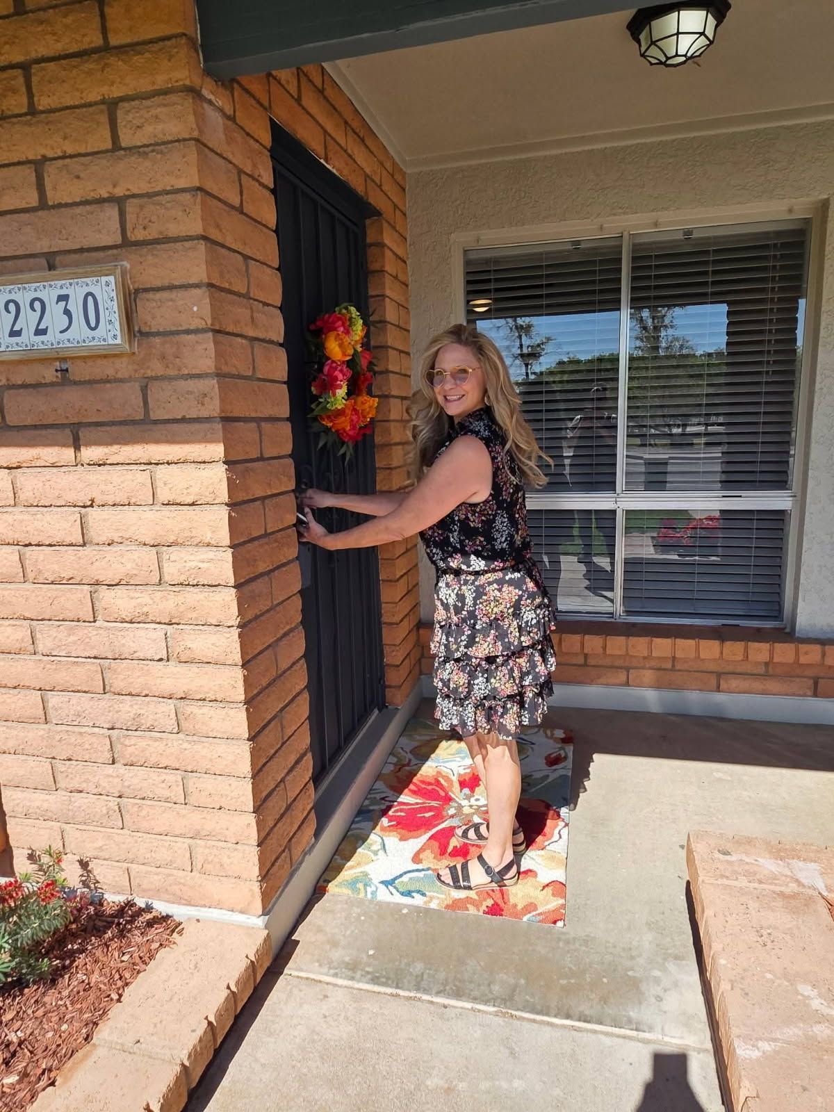 Woman at a front door on a brick porch holding a wreath, wearing a floral dress and heels