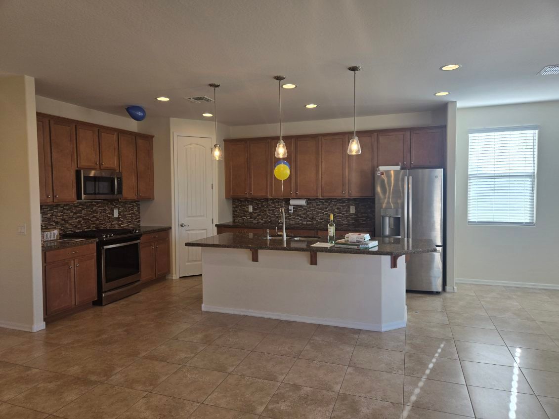Empty kitchen and dining area with wood cabinets, stainless steel appliances, island, and tiled floor.