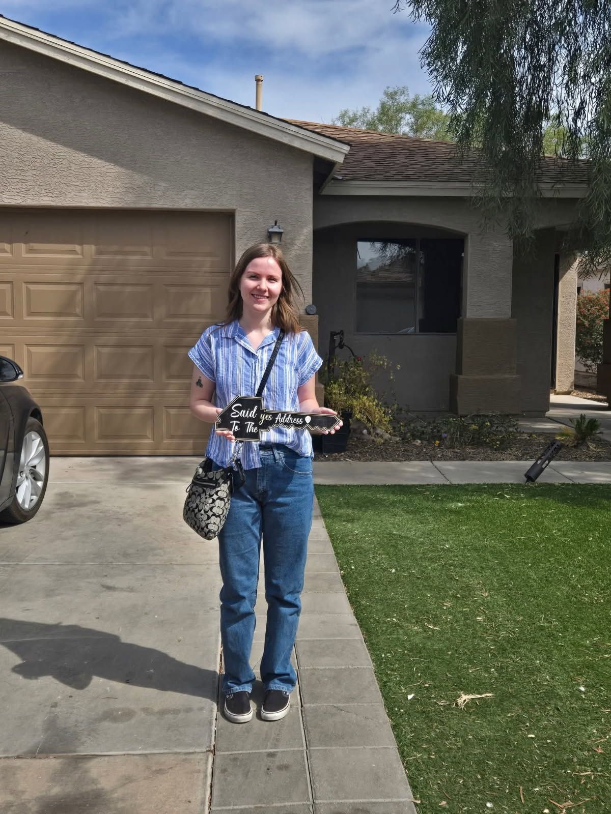 Person standing on a sidewalk in front of a house, holding a camera and smiling.