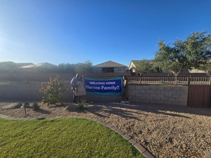 Blue banner on a front yard fence reads “Welcome Home,” with a house and bright sky behind it