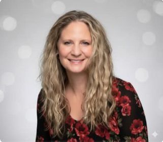 Smiling woman with wavy blonde hair wearing a black top with red floral pattern against a gray background