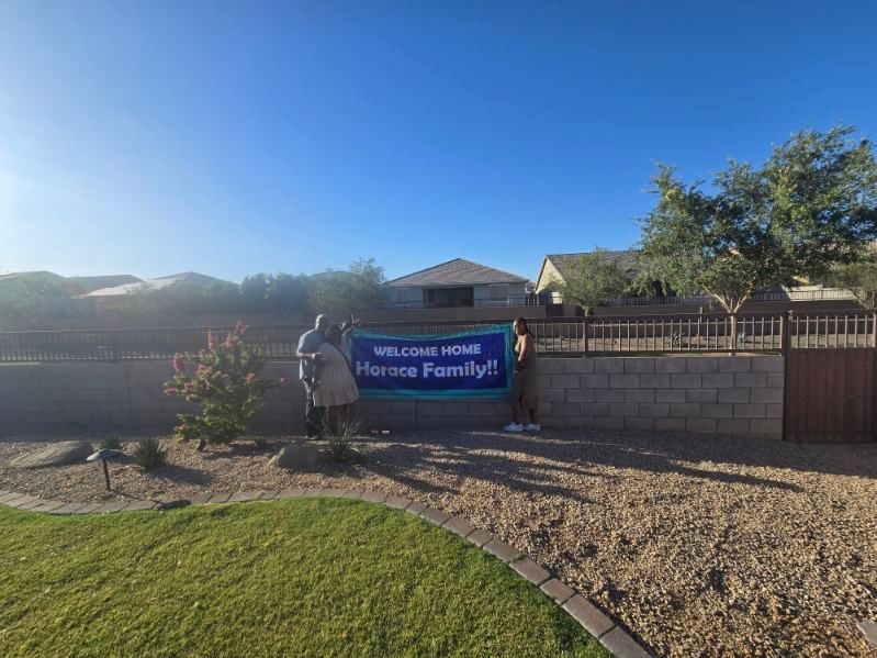 Neighborhood welcome sign reading “Welcome Home Family” beside a stone wall and desert landscaping under a clear blue sky