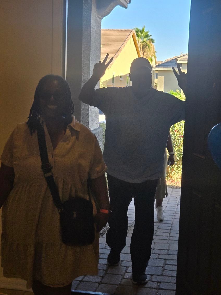 Two people standing in a doorway, one waving, with bright outdoor light behind them