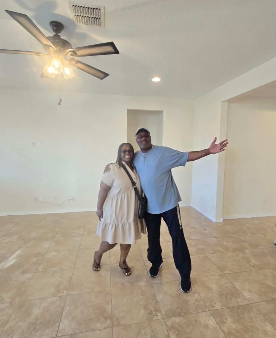 Two people posing in a mostly empty room with tiled floor and a ceiling fan.