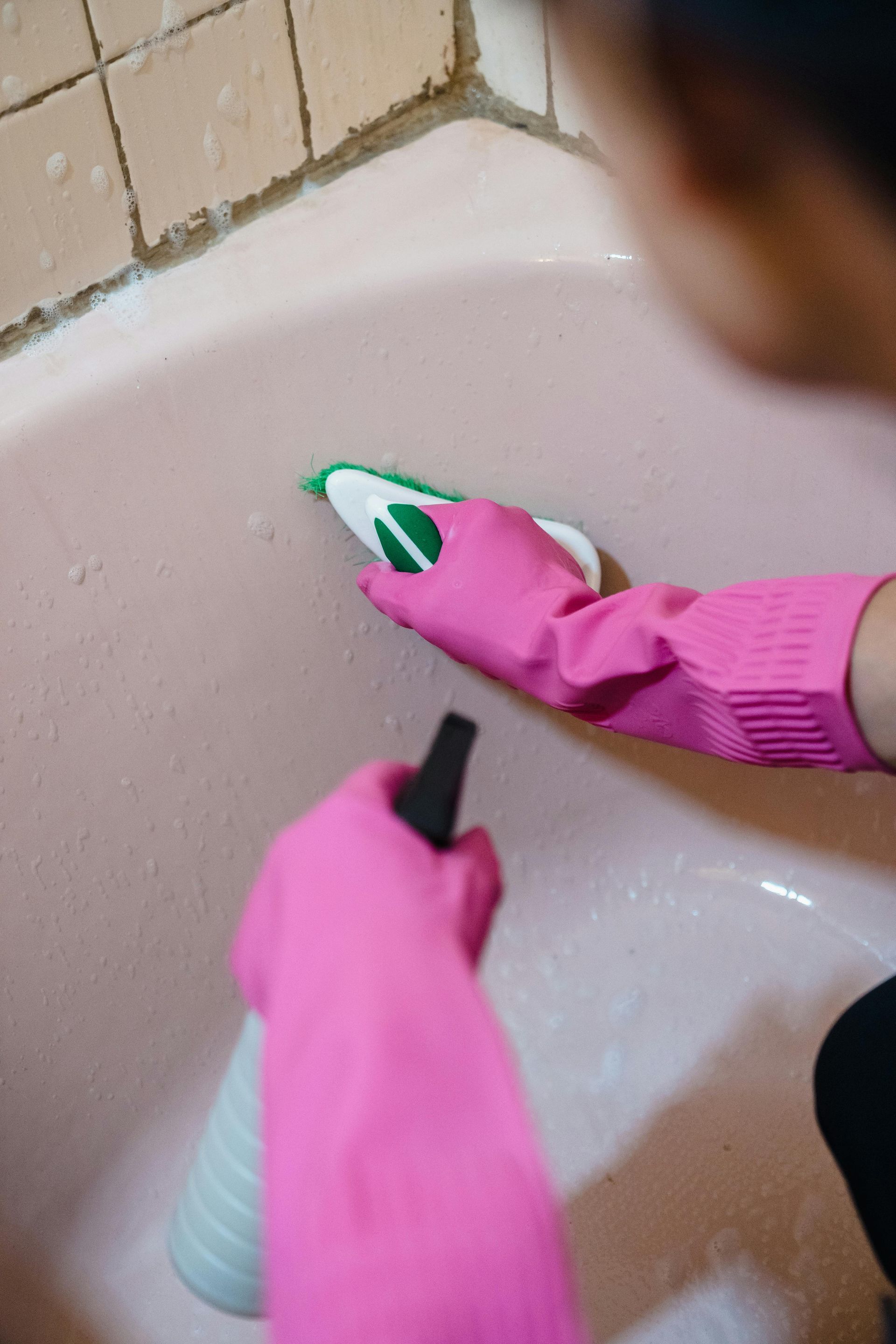 A person wearing pink gloves is cleaning a bathtub with a spray bottle.