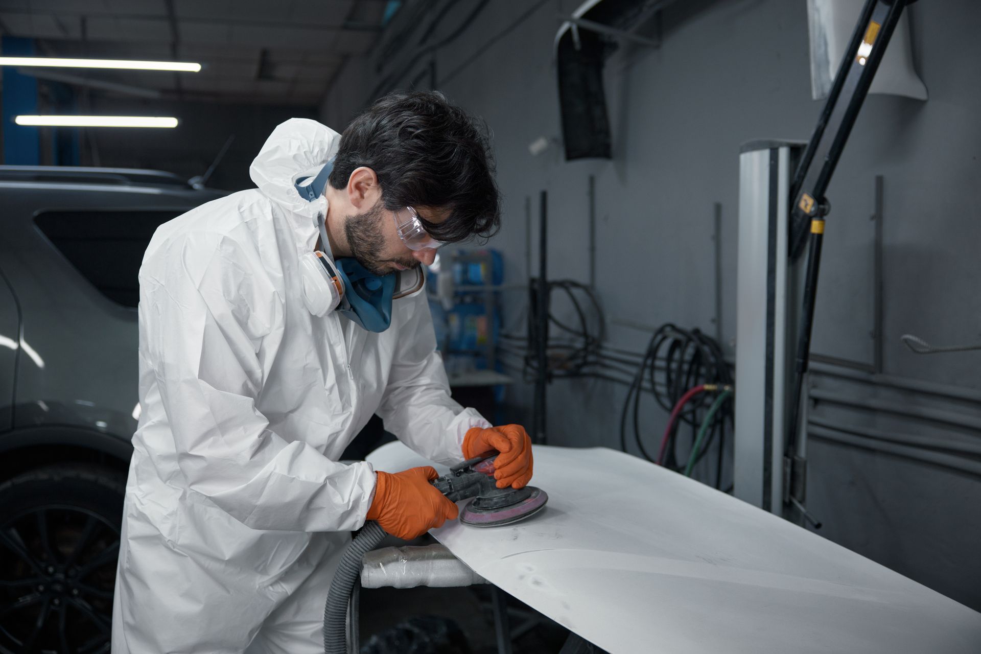 An auto body technician in a protective suit meticulously sands a car hood at a repair shop. An auto body technician in a protective suit meticulously sands a car hood at a repair shop.