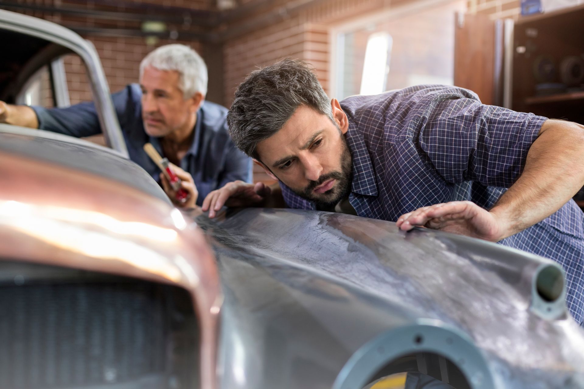Two focused mechanics examining a classic car panel in an auto repair shop. Two focused mechanics examining a classic car panel in an auto repair shop.
