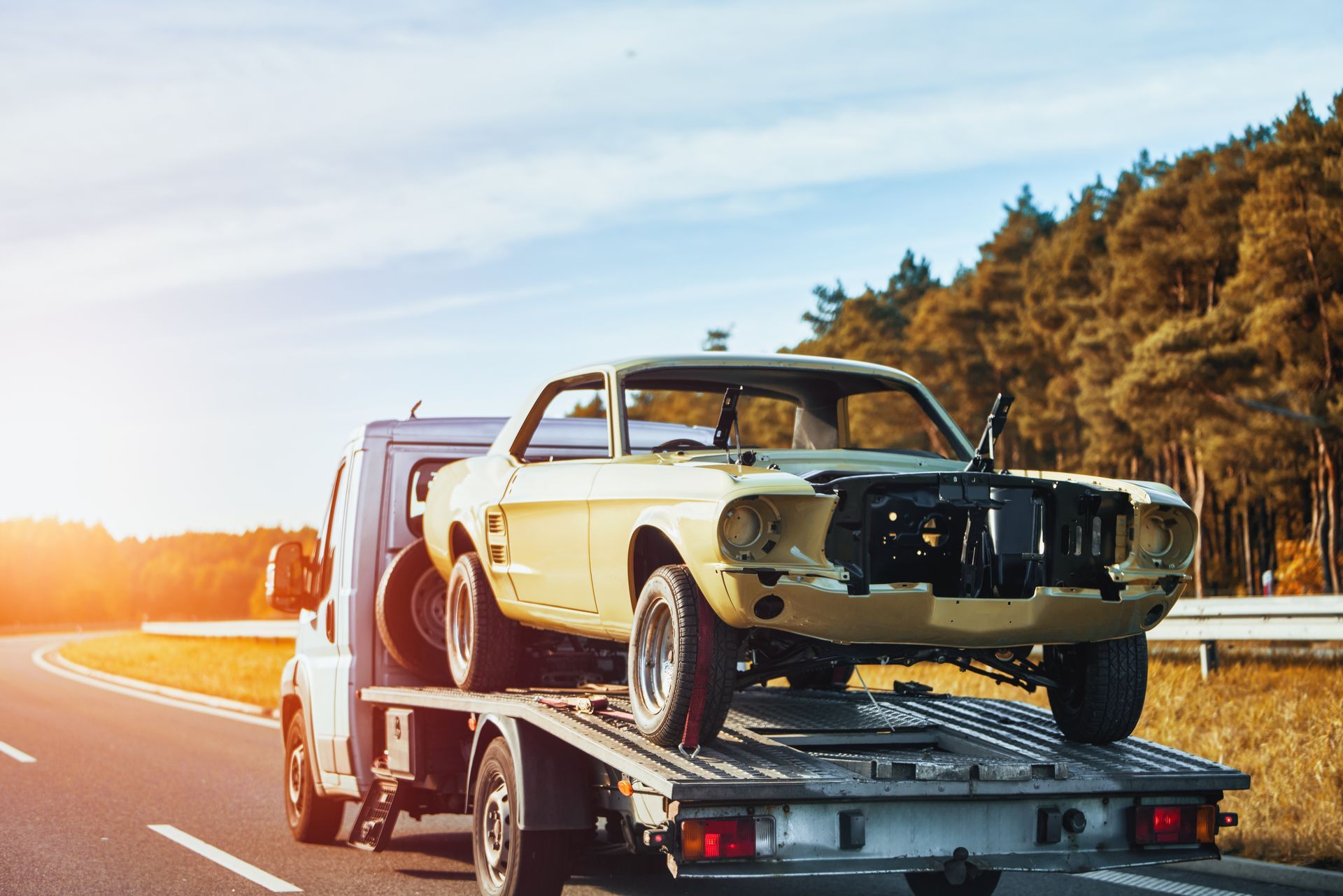 A tow truck in motion on a road with a damaged classic vehicle after being in a traffic accident.