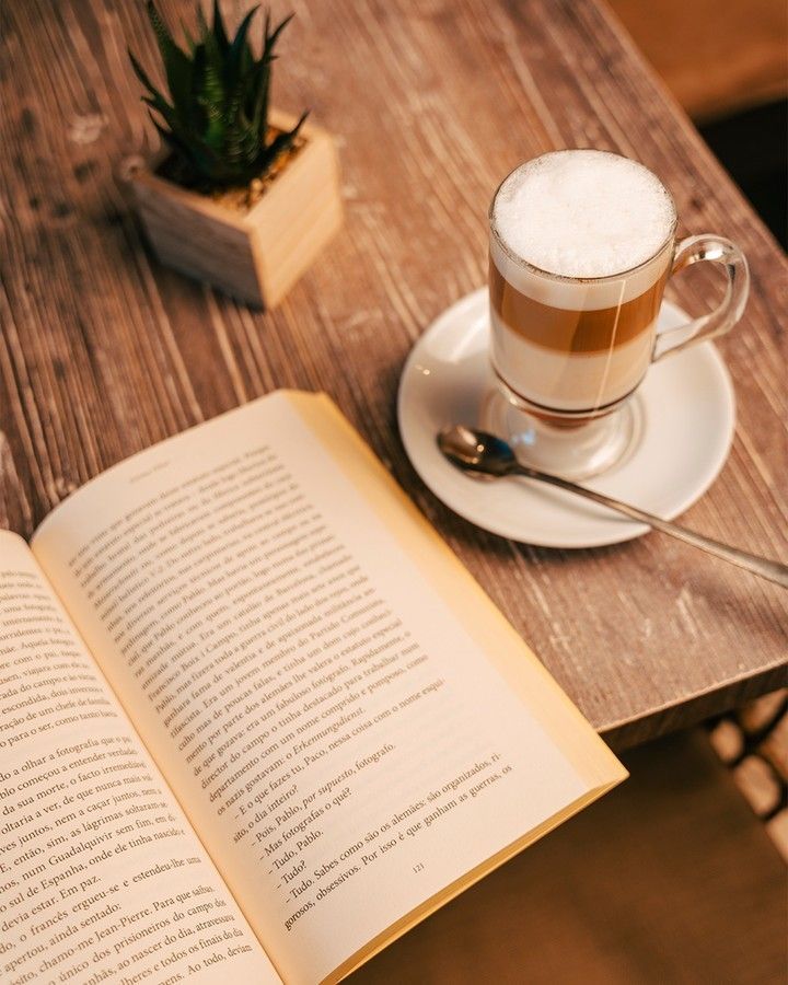 Uma xícara de café está em um pires ao lado de um livro aberto sobre uma mesa de madeira.