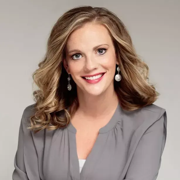 Portrait of a smiling woman in a gray blazer with wavy blonde hair and earrings