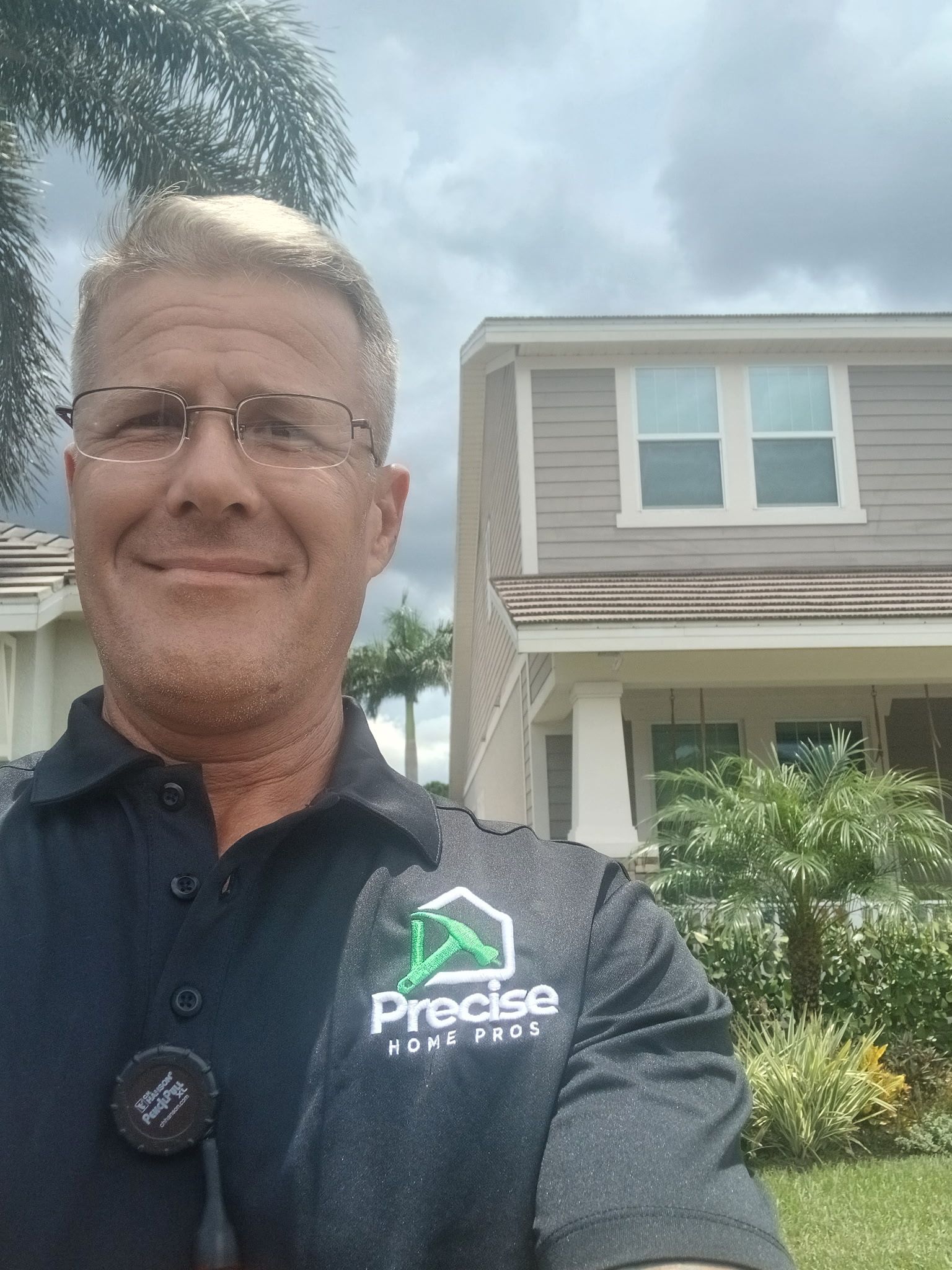 Man in dark polo shirt smiling in front of a house under a cloudy sky