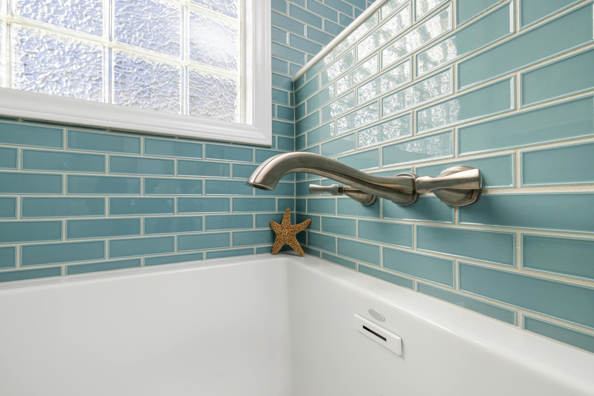 Bathtub corner with blue tile walls, frosted window, and wall-mounted faucet.