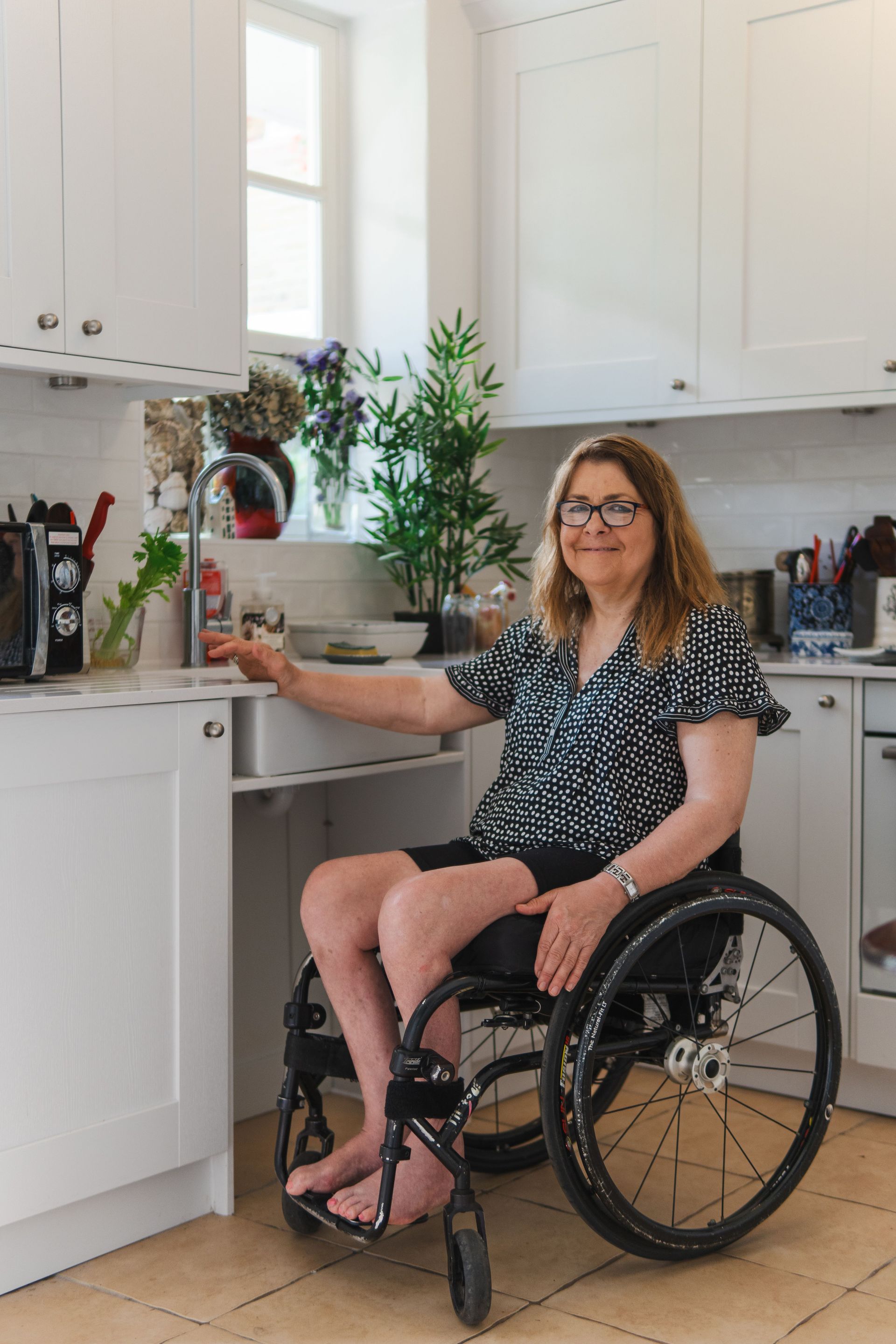Woman in a wheelchair in a bright kitchen, smiling beside white cabinets and a countertop with plants.