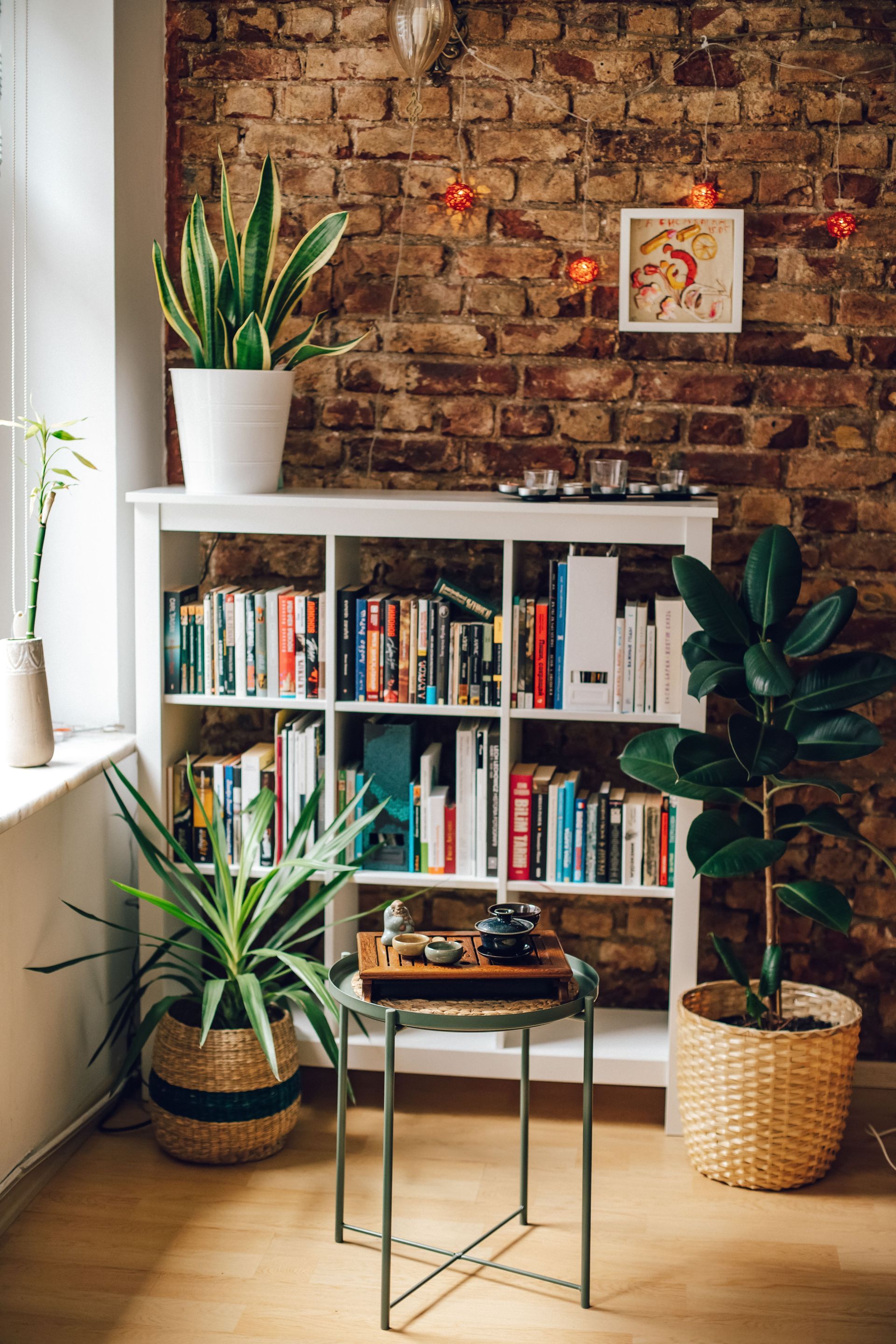 Cozy living room with a brick wall, white bookshelf, houseplants, and a small glass table by a window.