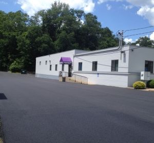 White single-story building with a purple awning beside a large empty parking lot under trees