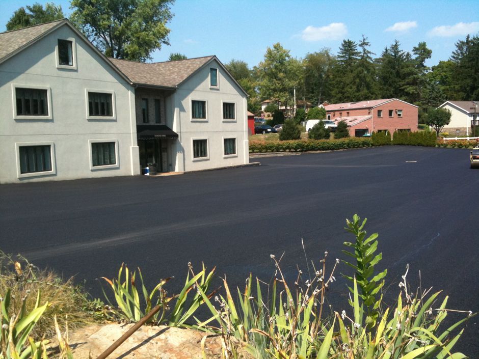 Large building beside a freshly paved blacktop lot with trees and a pink house in the background