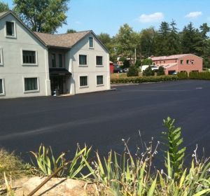 Two-story beige building beside a dark parking lot, with a pink building in the background and plants in the foreground