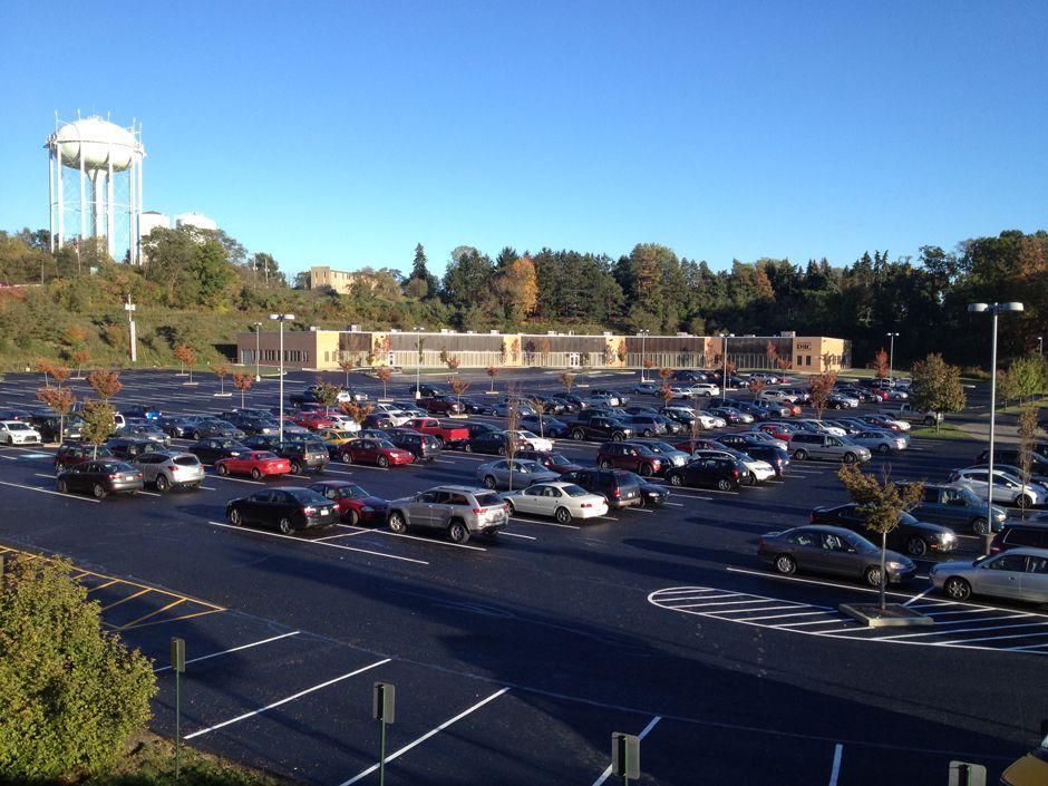Large parking lot full of cars beside a low building and trees, with a water tower in the background.
