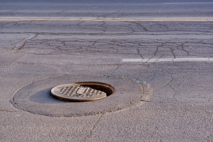 Open manhole in cracked asphalt road with metal cover slightly ajar