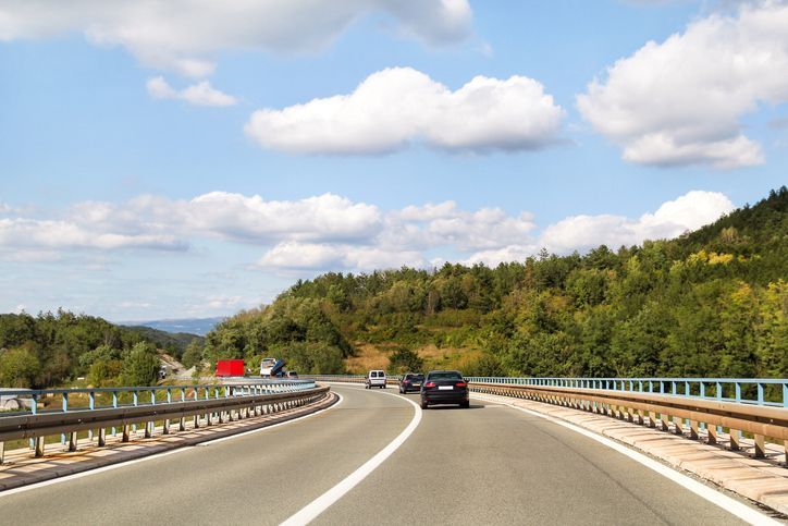 Curving highway through green hills under a blue sky with clouds, with a few cars ahead
