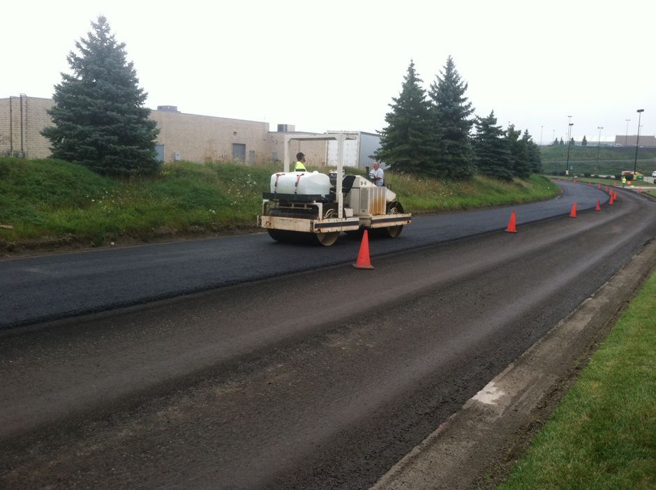 Roadwork vehicle paving a wet road beside orange cones and evergreens under overcast skies