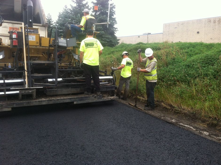 Road workers in reflective gear paving a street beside a truck and grassy embankment.