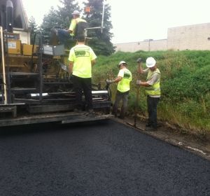 Road crew in neon safety vests working beside a paving machine on a wet road