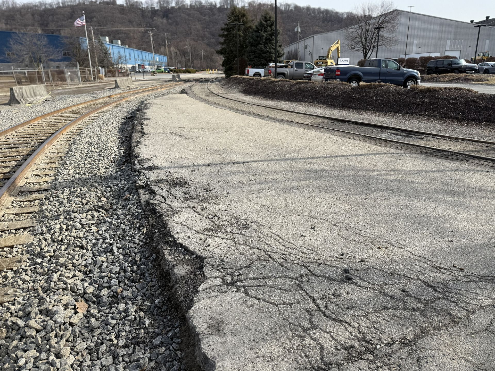Cracked, eroded roadside beside railroad tracks near industrial buildings on a gray day