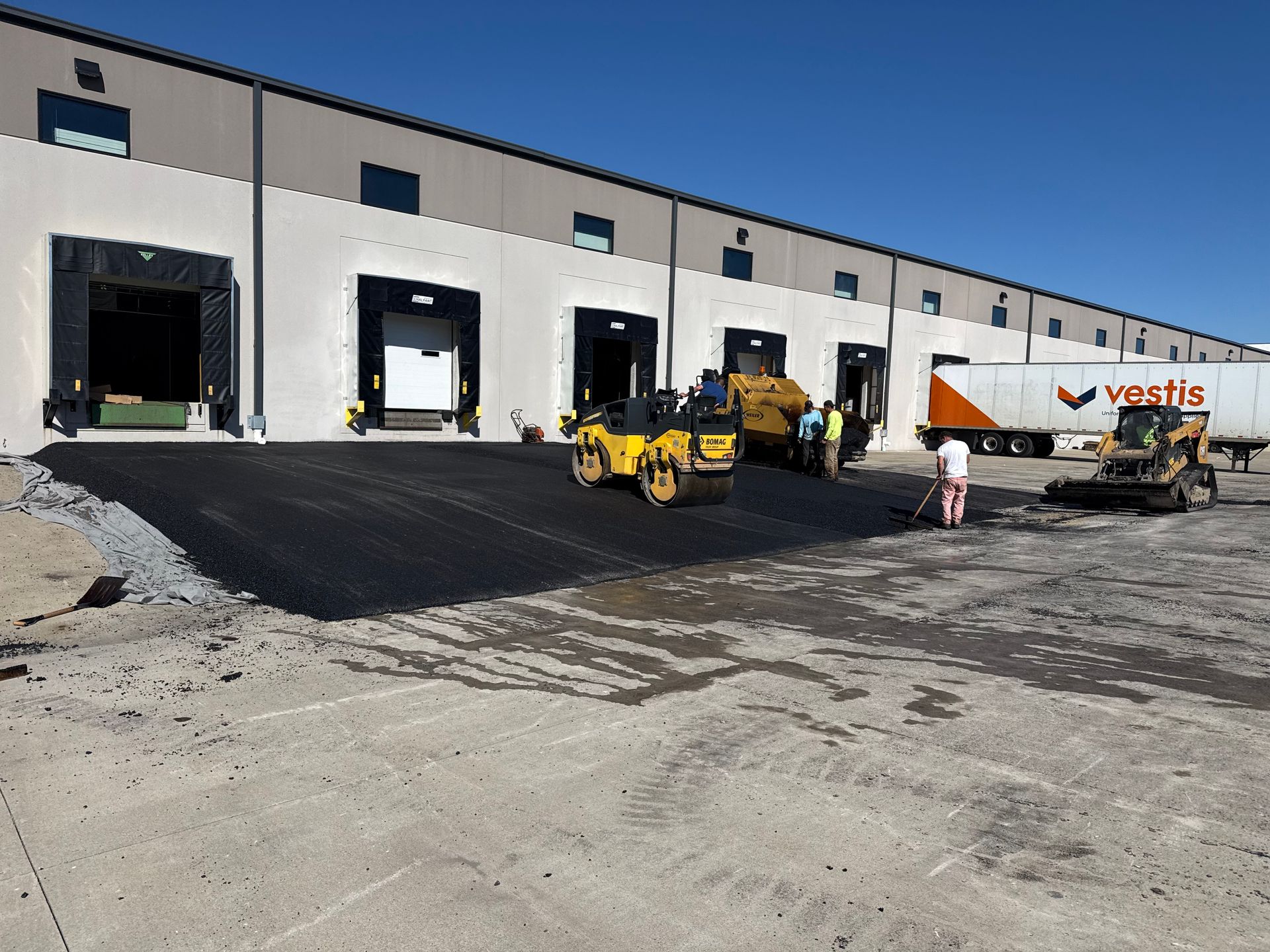 Construction crew paving a warehouse loading dock with asphalt and machinery under a clear blue sky