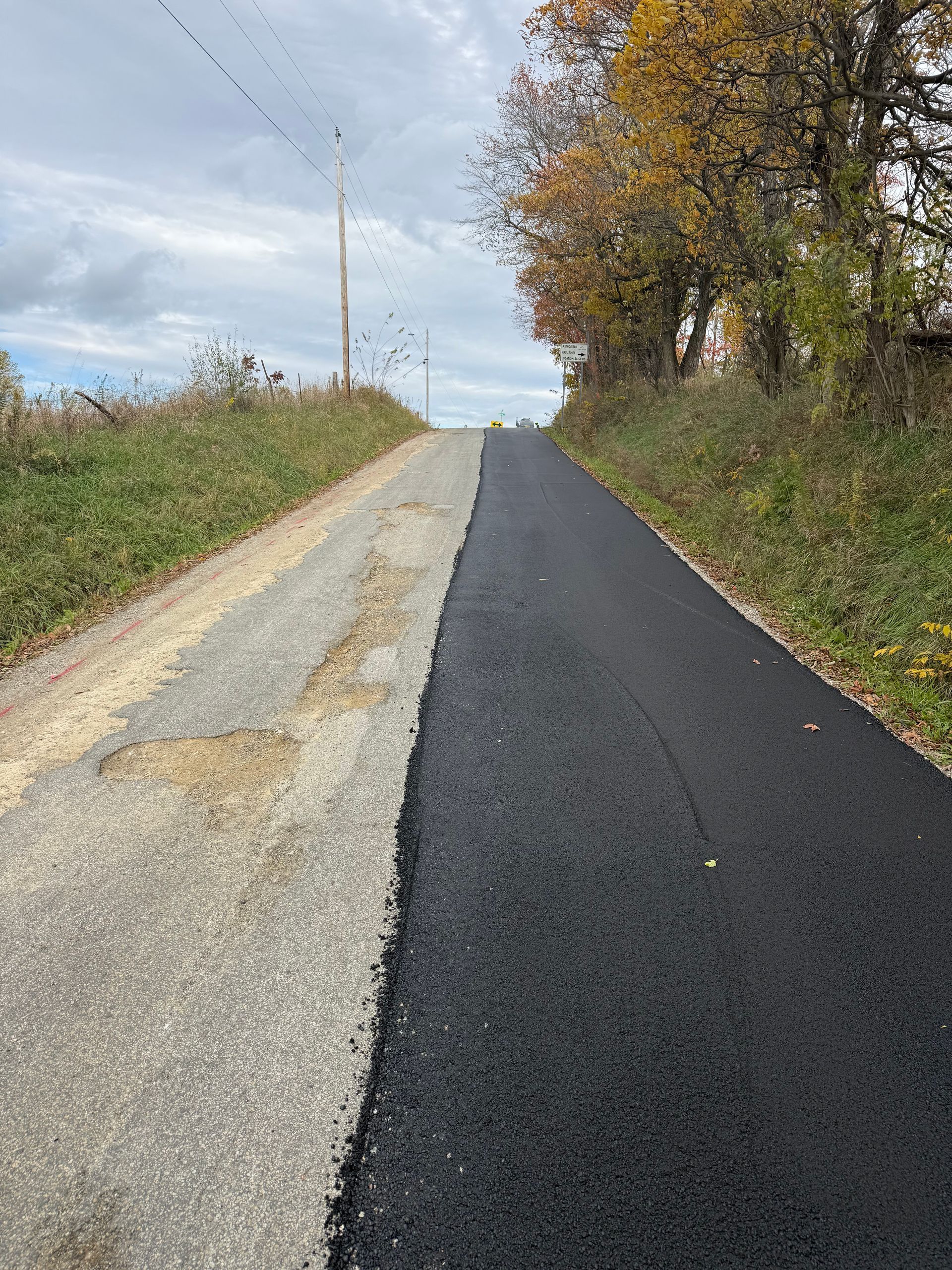 Fresh blacktop road beside a gravel shoulder and autumn trees under a cloudy sky