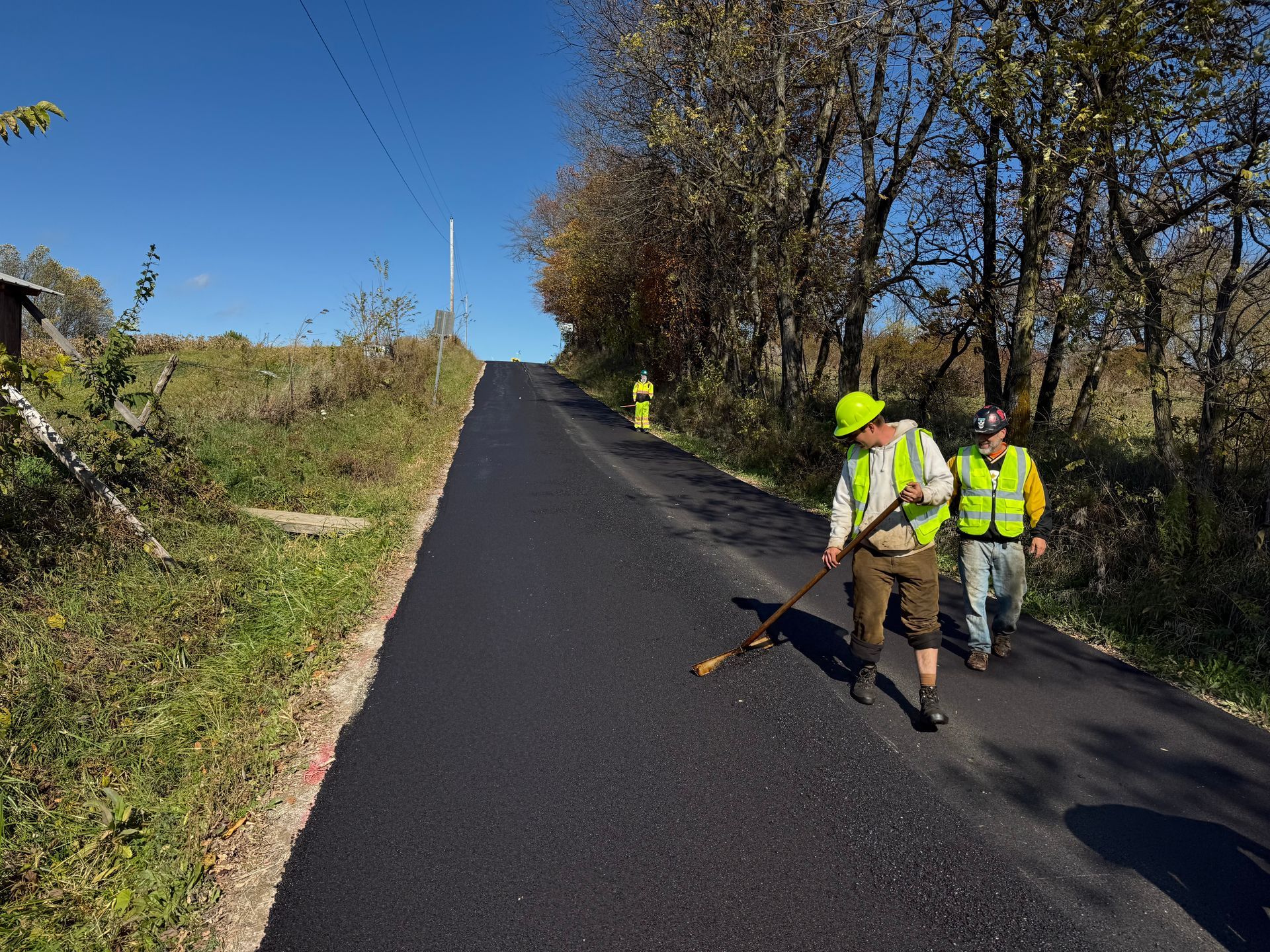 Road workers in neon vests repair fresh black asphalt on a rural road under blue sky.
