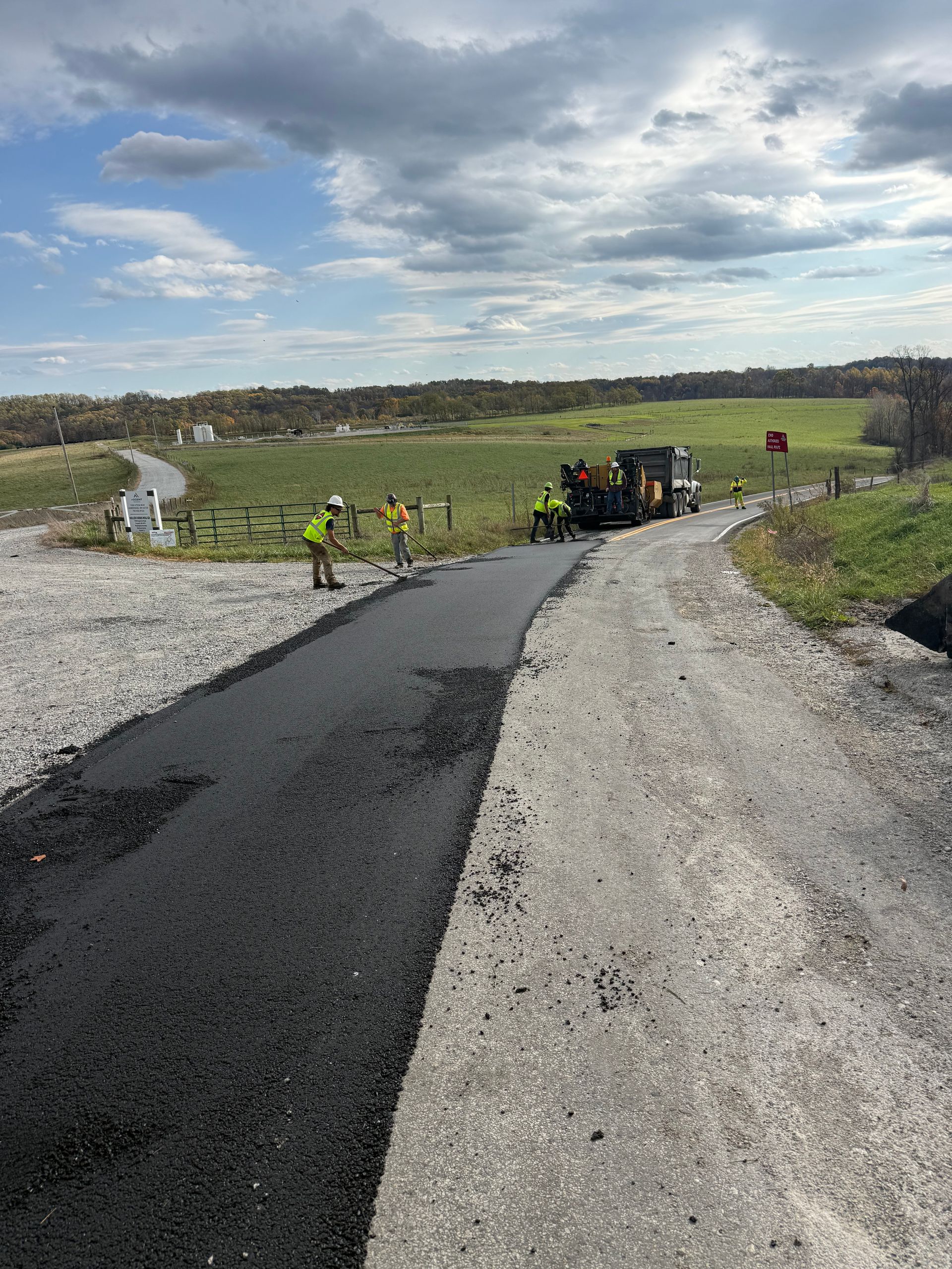 Workers paving a gravel road through a green field under cloudy skies