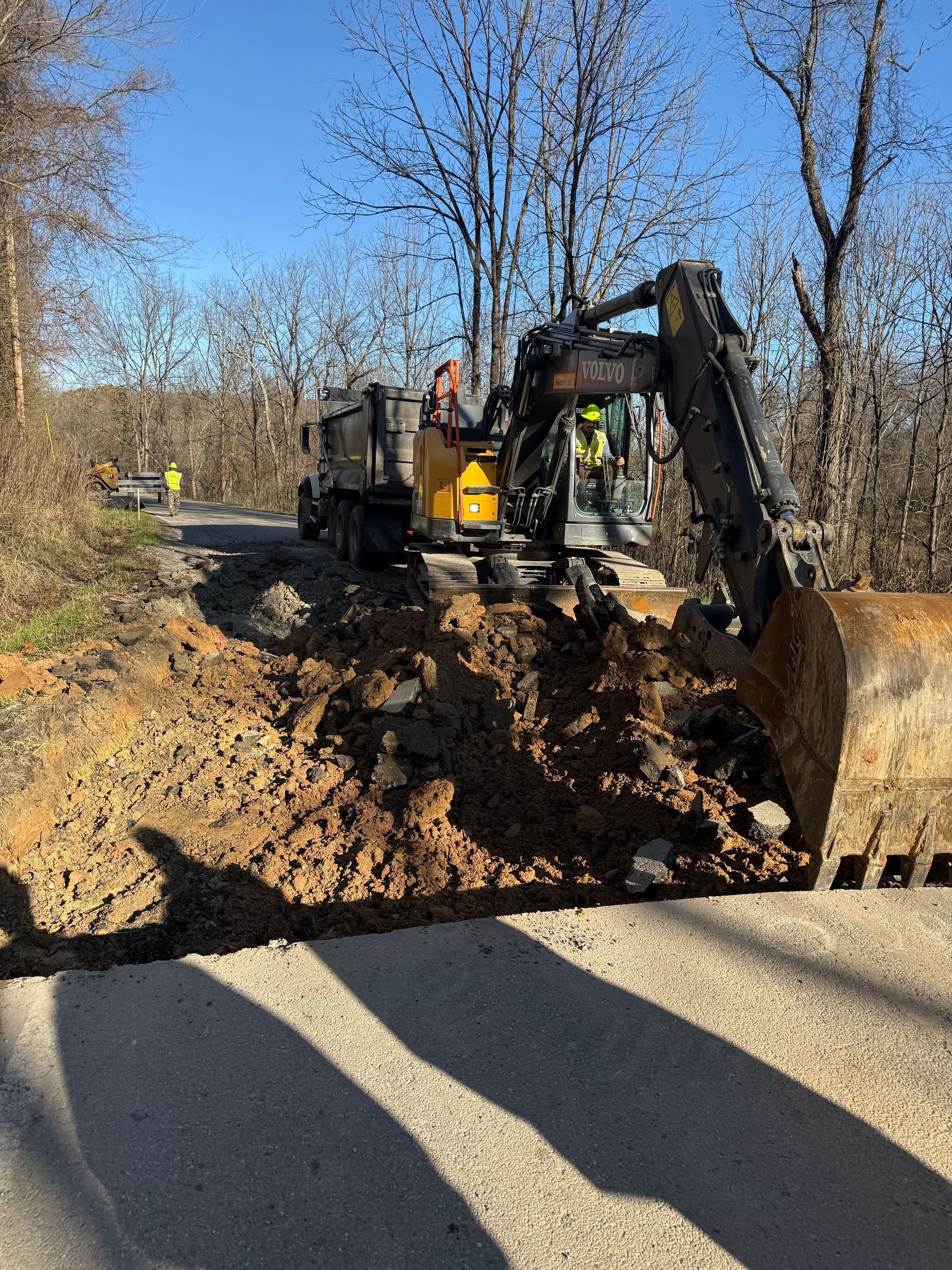 Backhoe clearing mud across a road on a sunny day, with a worker nearby and bare trees lining the roadside