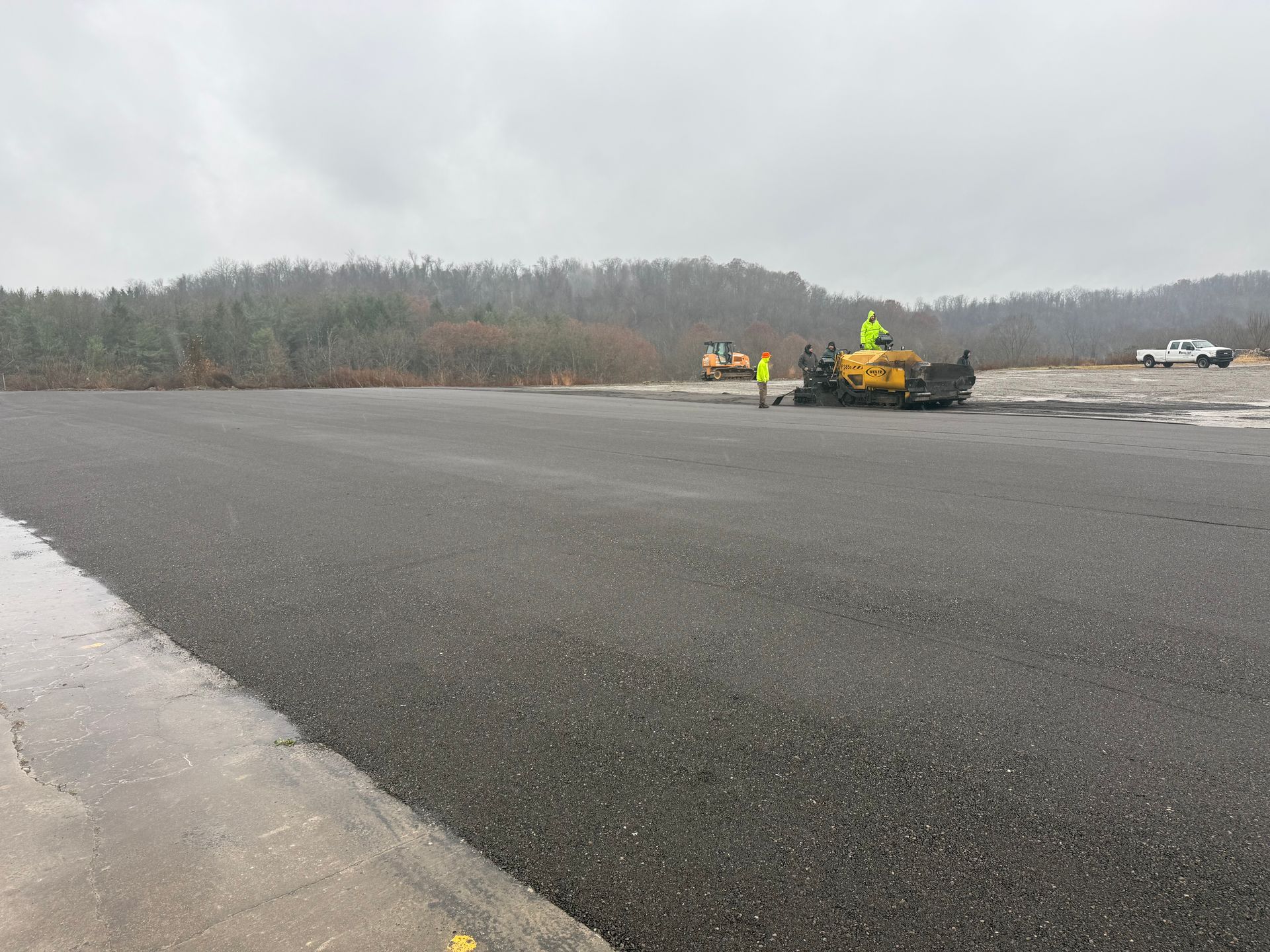 Workers and yellow equipment paving a wide gray road beside a wooded area