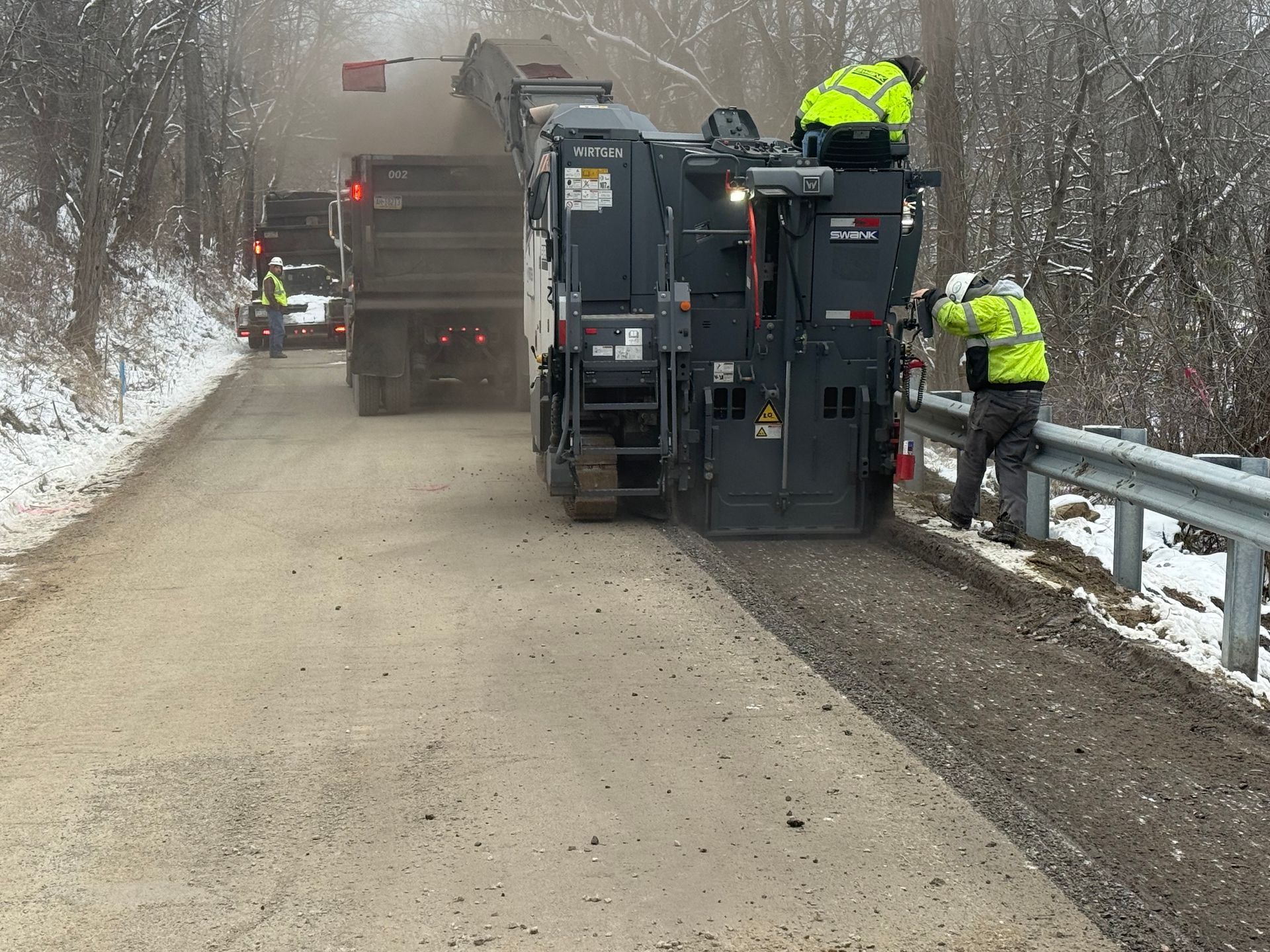 Road crew in neon jackets repairing a muddy rural road beside a guardrail in winter weather