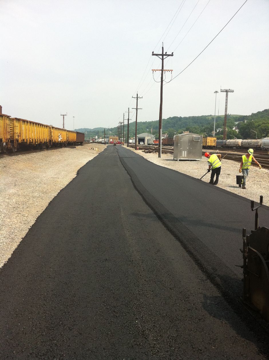 New black asphalt road under construction beside railroad tracks, with workers and utility poles.